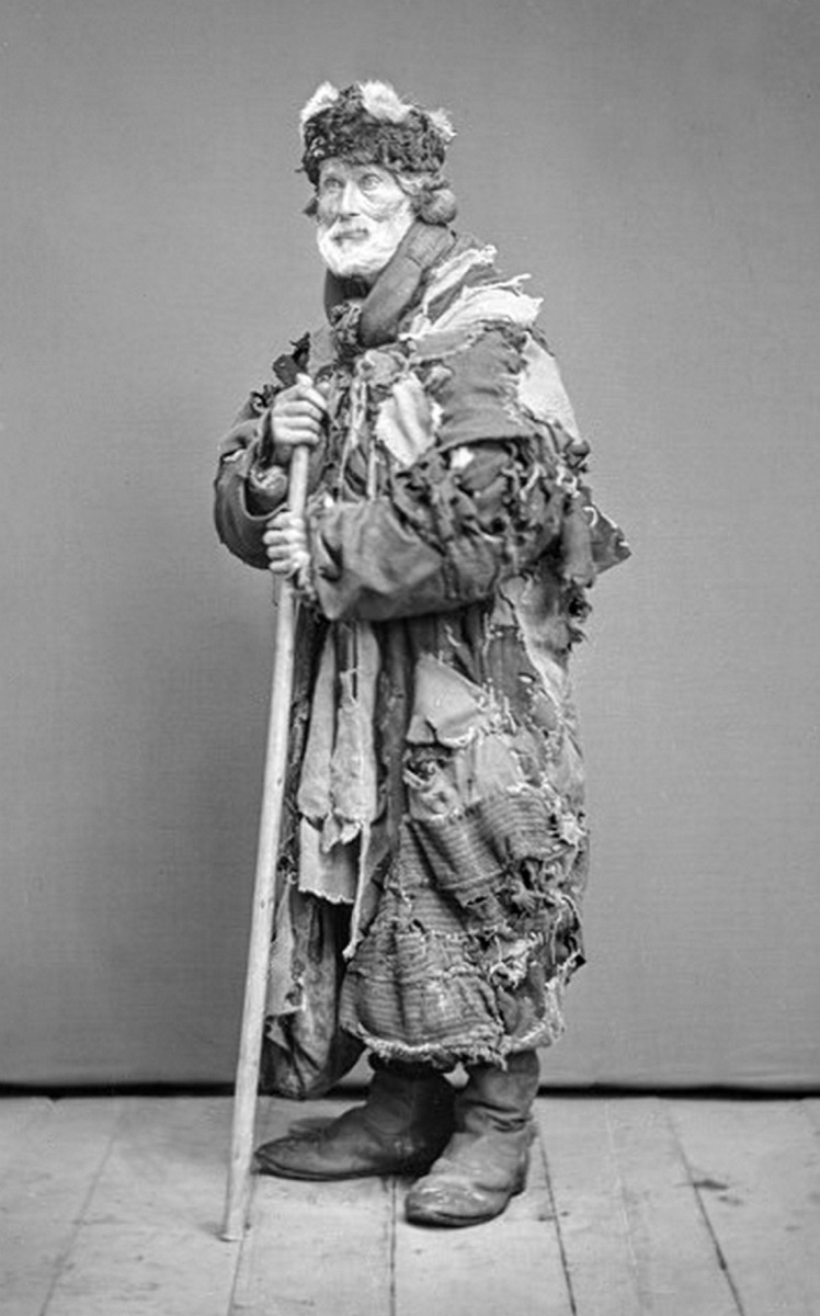 A full-length portrait. An elderly man with a grey beard, in a fur hat and a long patched coat, leans on a stick with both hands.