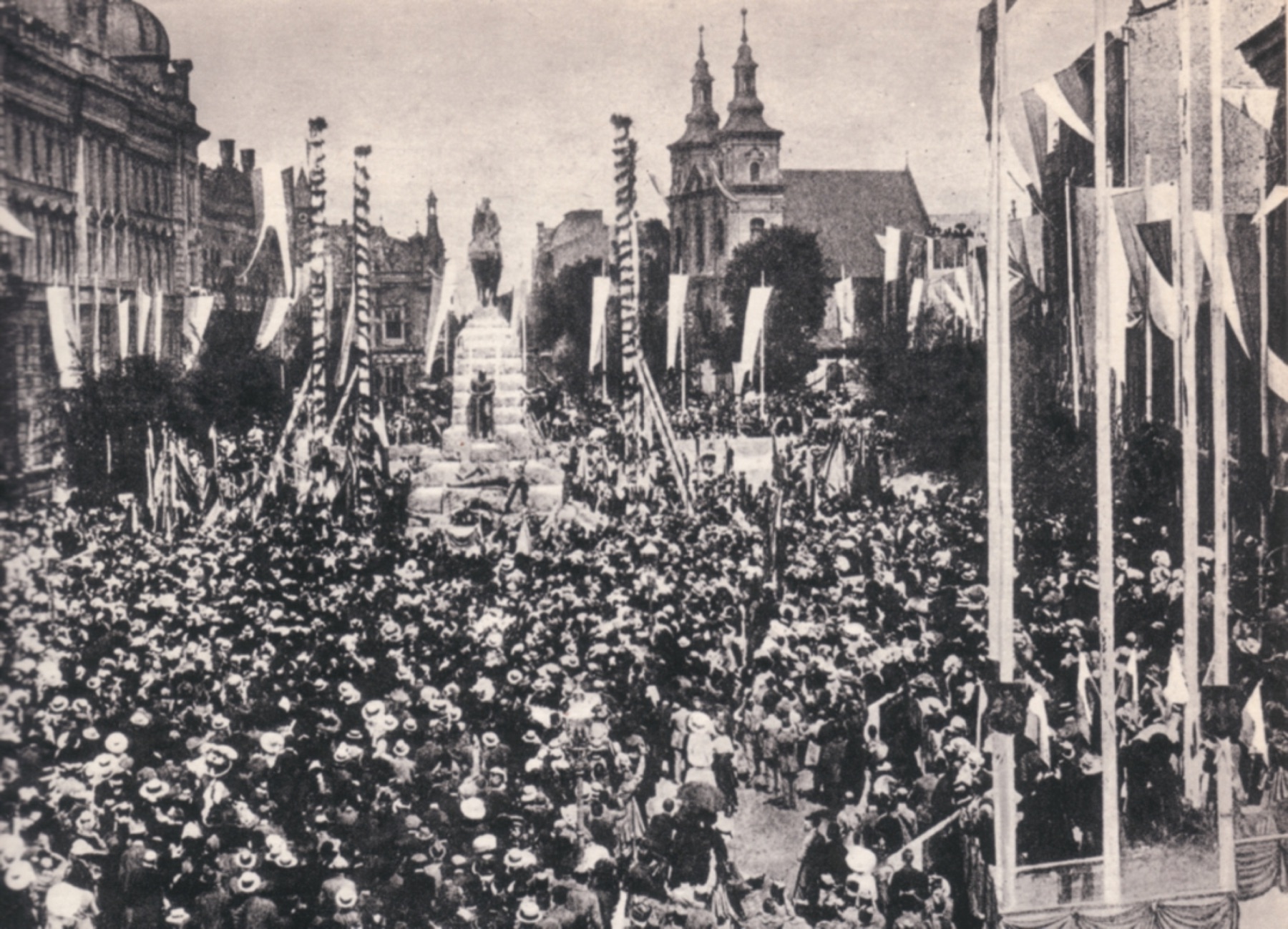 A black-and-white photograph from 1910. A crowd of people in hats fills the square, with flags and tall masts wrapped in garlands above them. In the background, on a stepped pedestal, an equestrian statue of the king, and behind it the two towers of St Florian's Church. On the left, a large building with a dome.