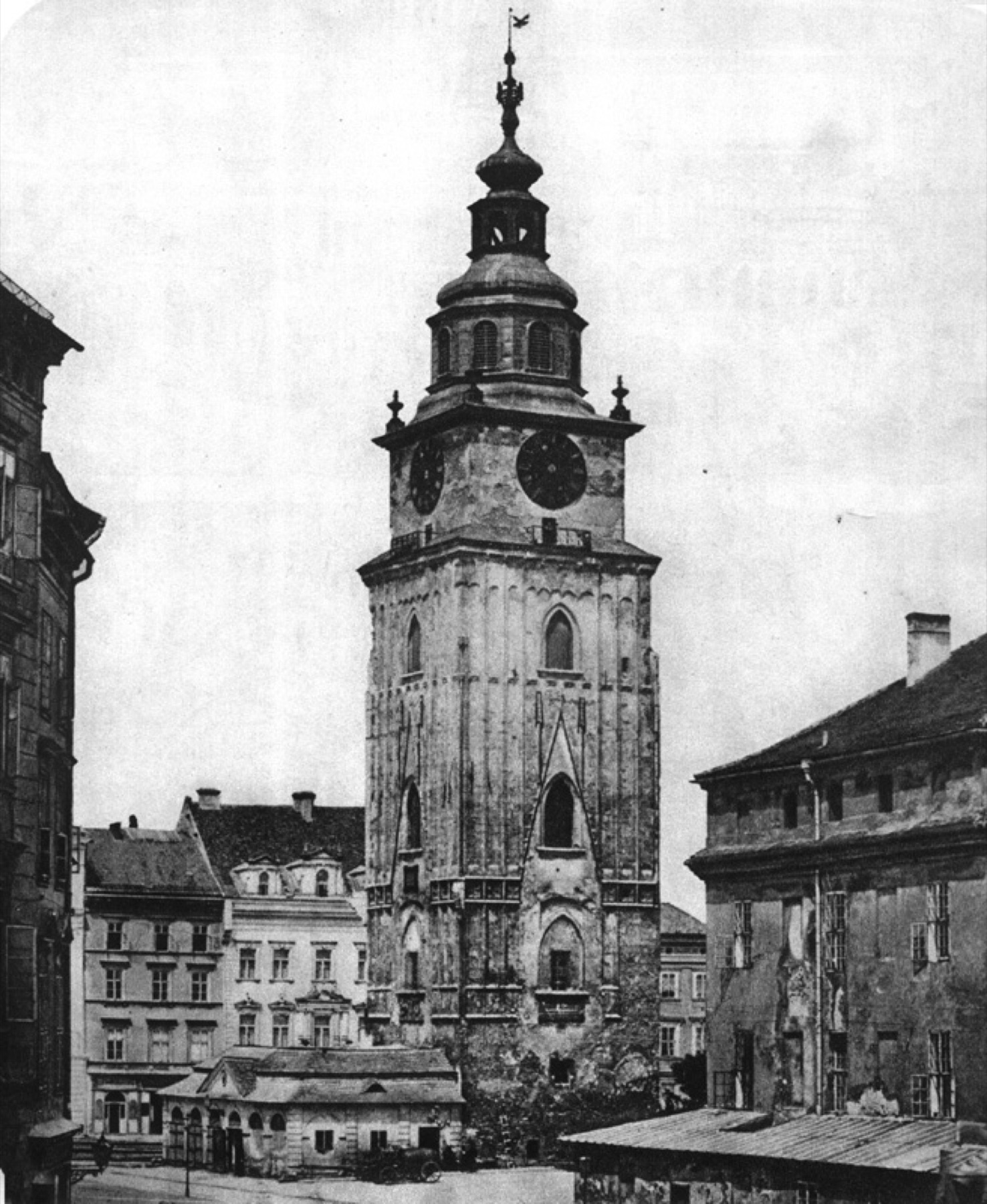 A tall stone tower with Gothic windows and a clock, topped by a bulbous cupola. At its foot, a low building; on the right, the dark wall of the building next door.